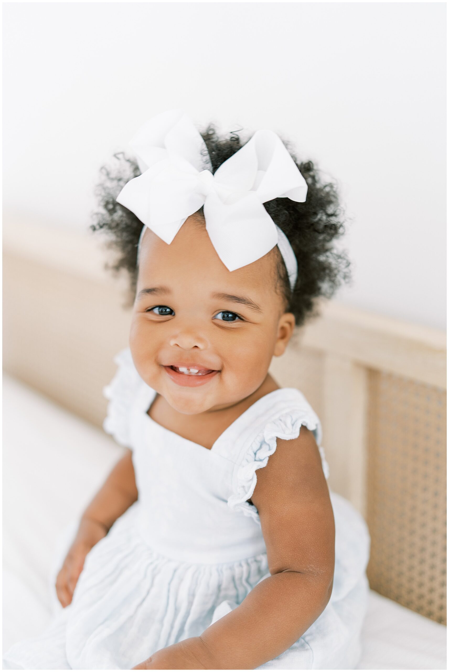 Portrait of a one year old girl in a white dress with a large white bow on her headband.