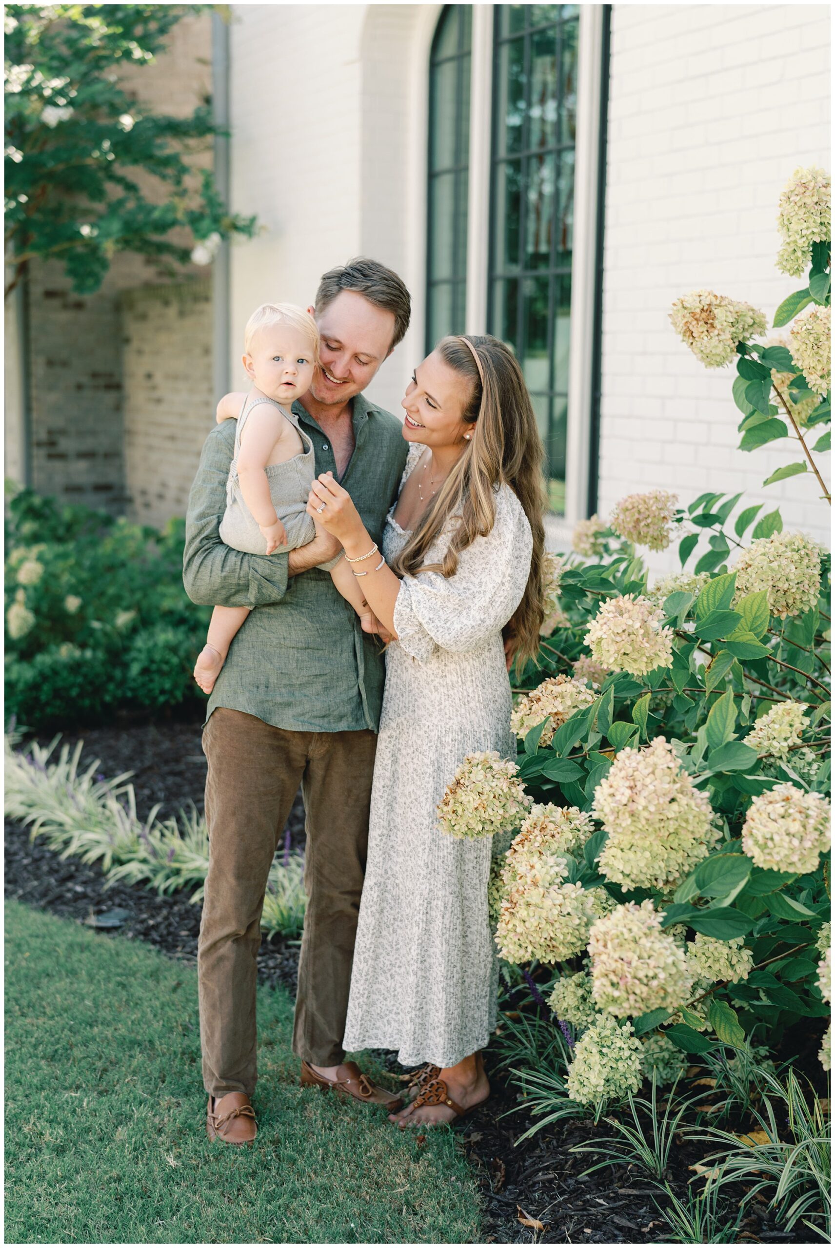 Parents pose smiling at their one year old boy in front of a white brick home with lush green hydrangeas surrounding them for an Alpharetta family photography session.