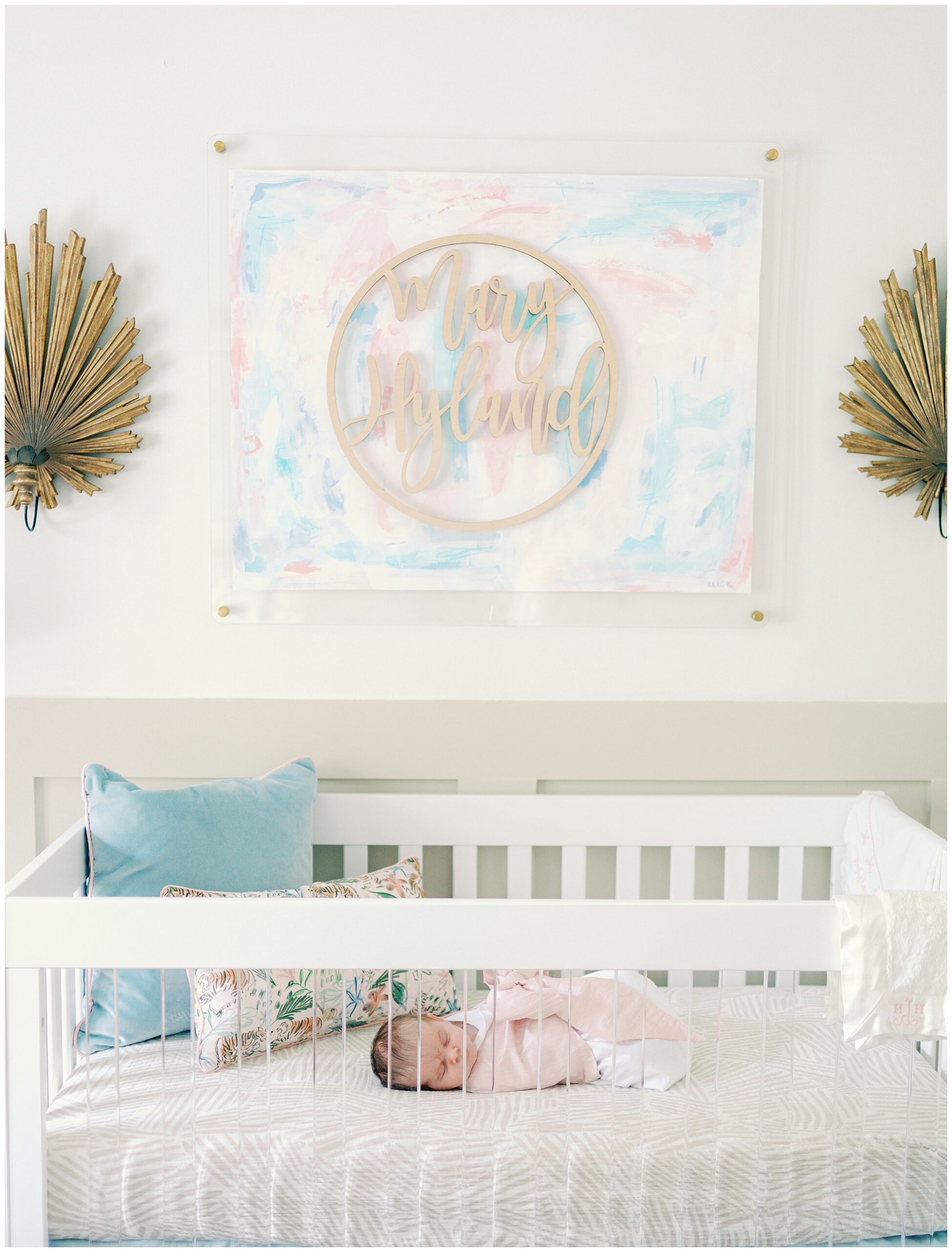 Image of classic girl nursery decor with a newborn baby laying in a white crib with acrylic front and a watercolor name painting hanging on the wall flanked by two gold palm frond wall art pieces.