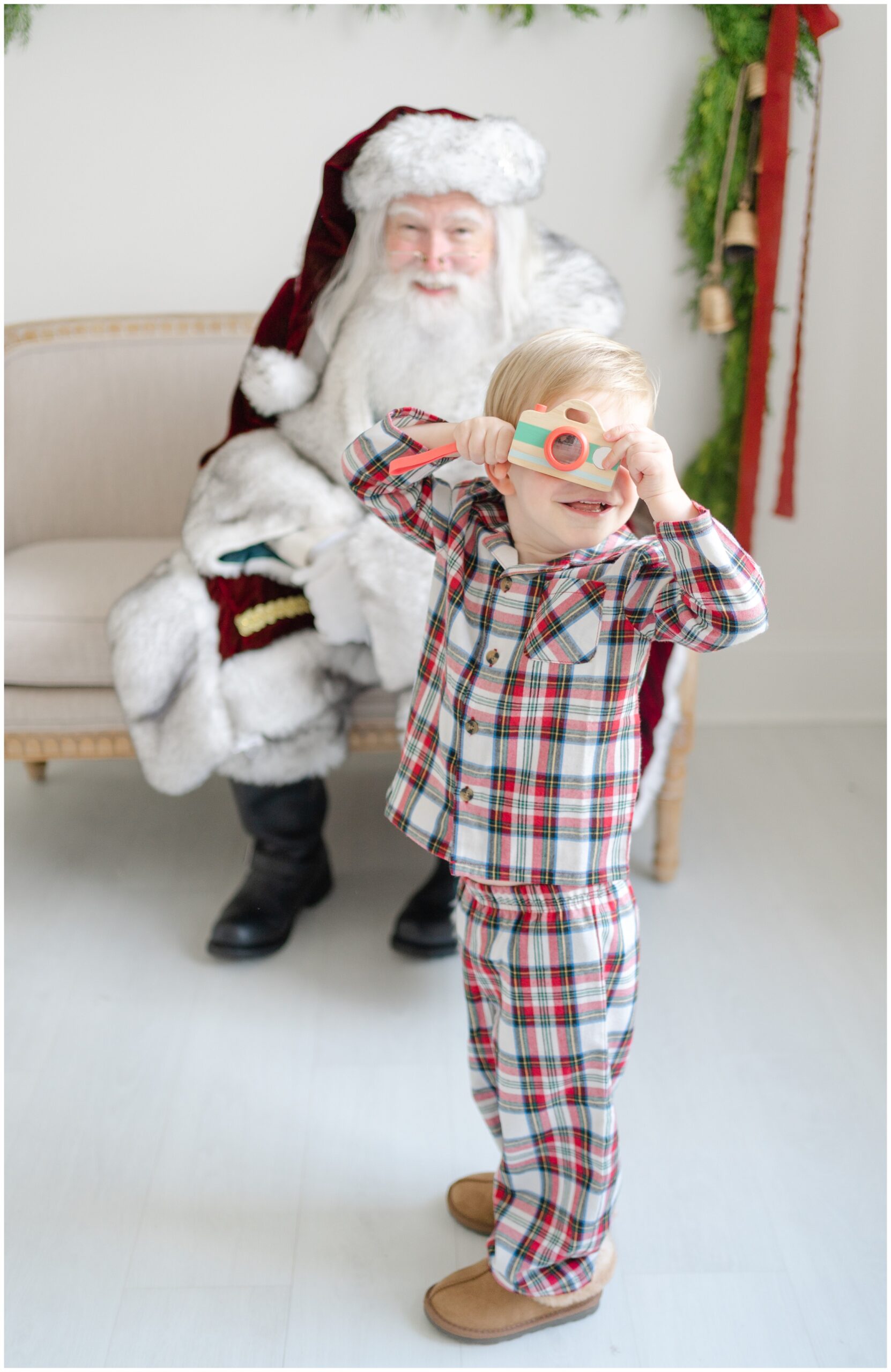A young boy wearing plaid Christmas pajamas holds a camera to his eye while standing in front of Santa. 