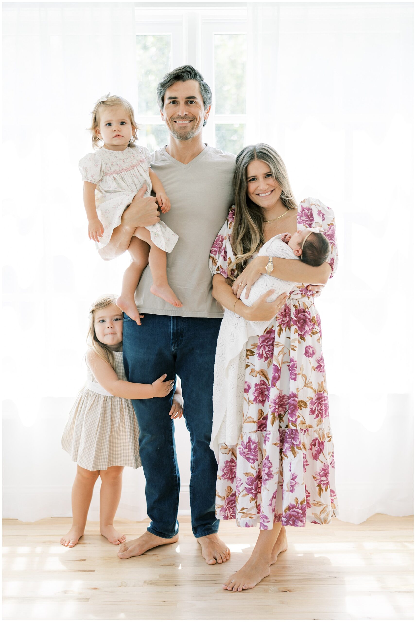 Parents standing in front of a window with white curtains holding a newborn baby girl and a toddler daughter with their older daughter standing and holding onto her father's leg for a Roswell newborn photo session.
