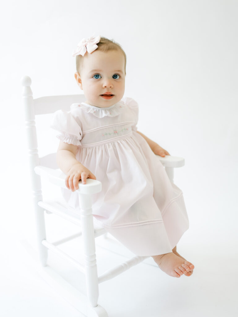 Little girl sitting in a white rocking chair during a portrait session with Atlanta photographer, Grace Emily Photography. She is wearing a pink bow & a pink dress