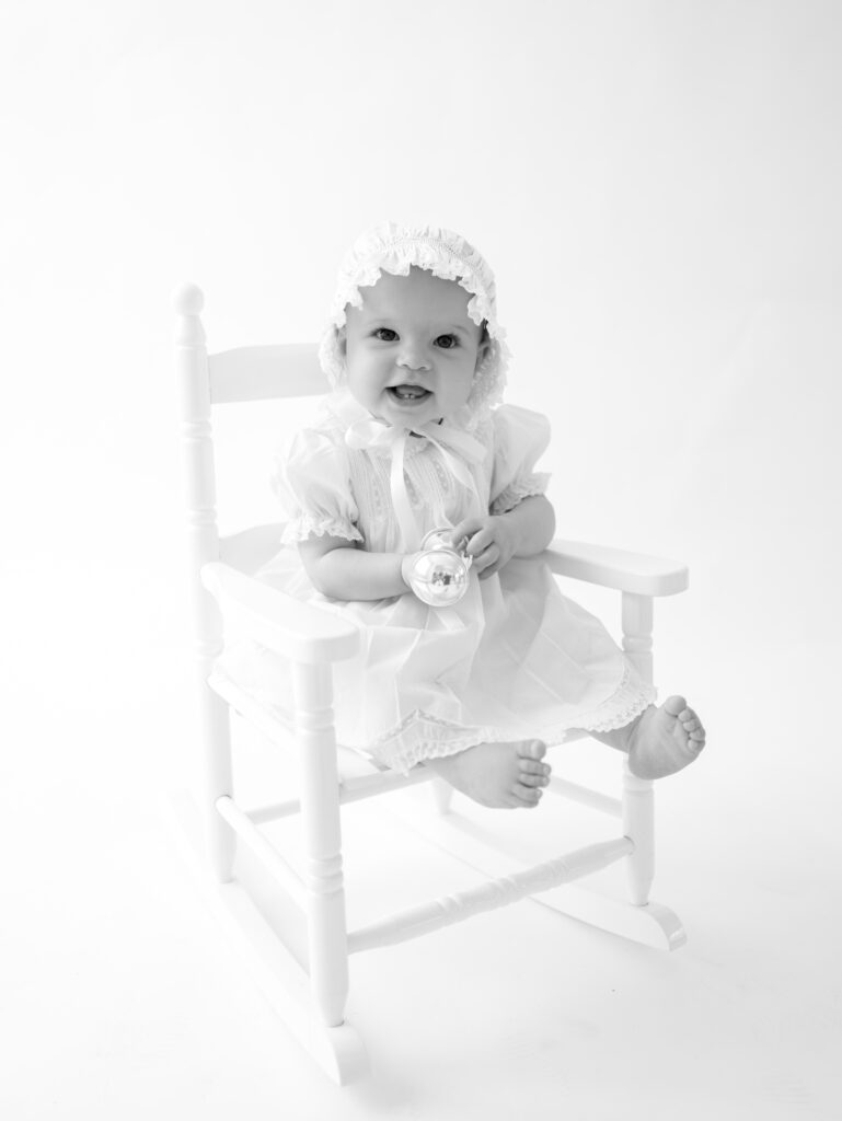 Little girl sitting in a white rocking chair, holding a silver rattle chair during a portrait session with Atlanta photographer, Grace Emily Photography.