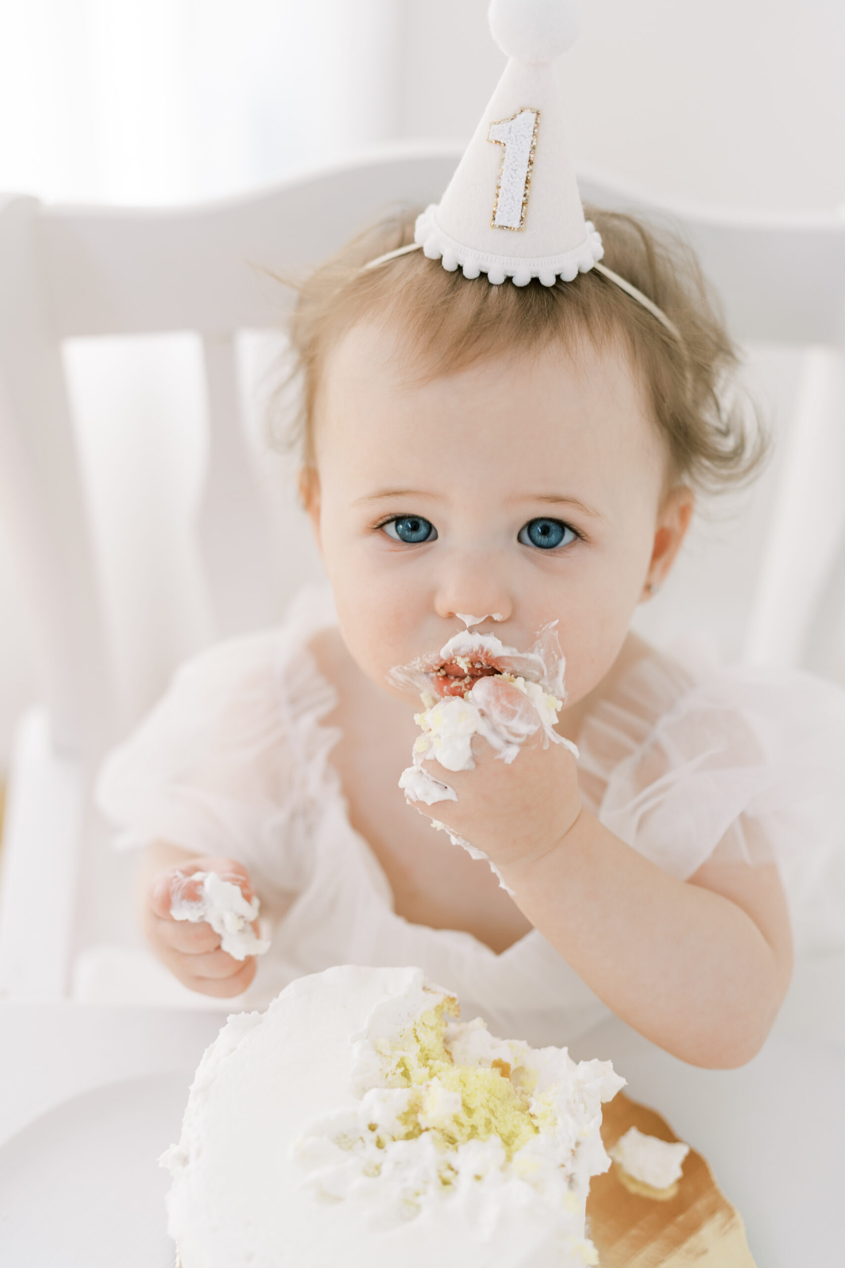 A little girl with blonde hair wearing a white birthday hat eating a vanilla cake.