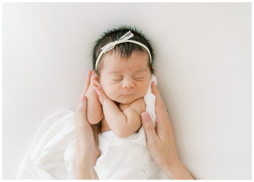 A picture of a newborn baby with dark black hair wearing a tiny white lace bow. She is having her picture taken by Roswell newborn photographer, Grace Emily Photography. 