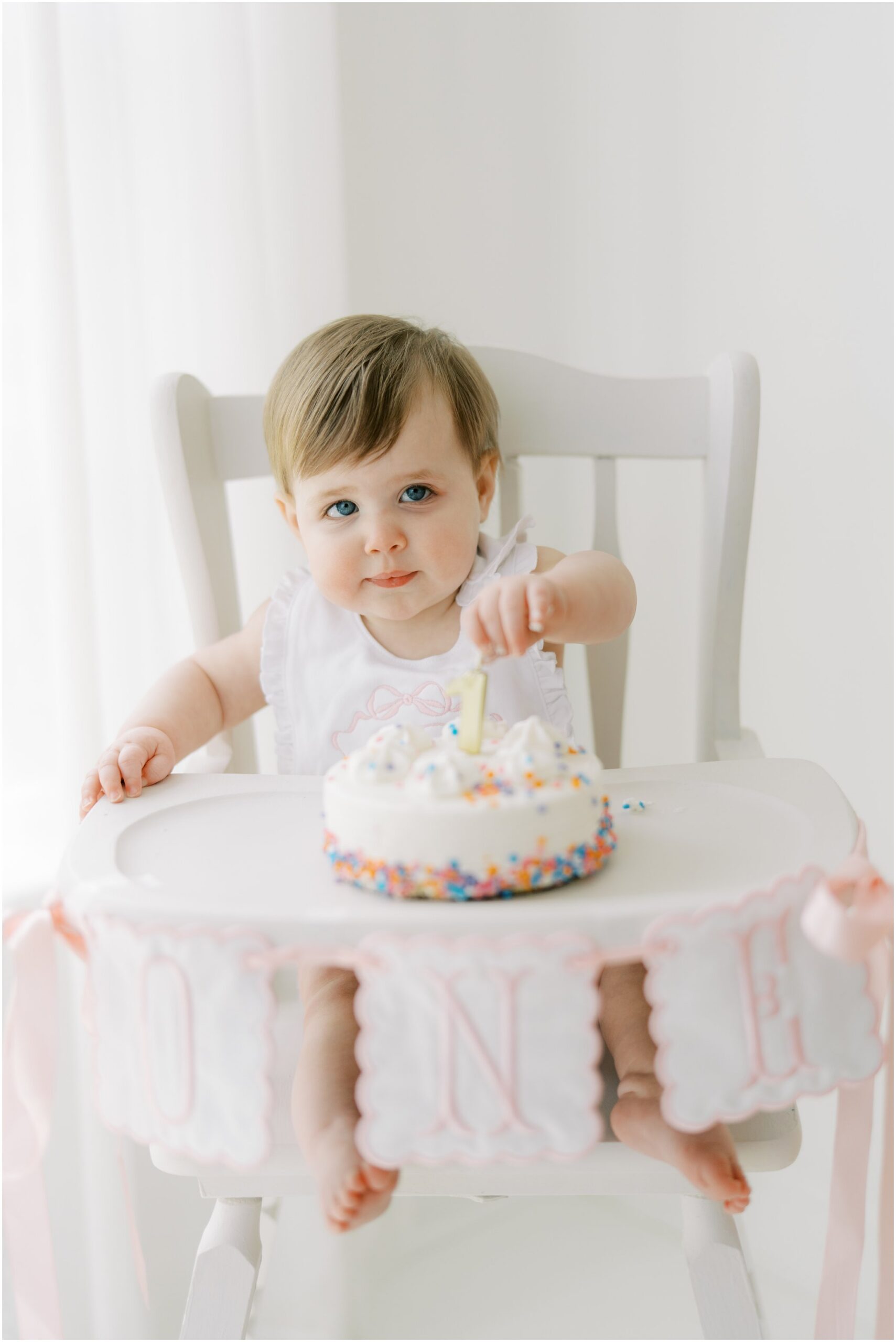 Little girl sitting in a white high chair eating a white birthday cake